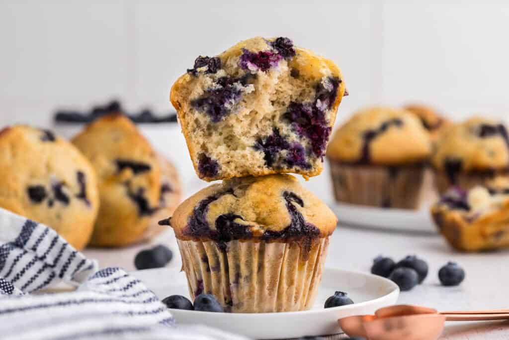 Side shot of a vertically cut open blueberry cottage cheese muffin stacked on top of another whole muffin. Inside there are lots of dark blue blueberries present. More muffins are in the background.