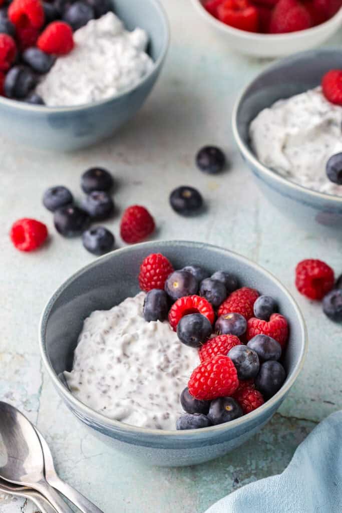 Low angle shot of a Greek Yogurt Protein bowl. The small bowl is filled with creamy white yogurt mixed with chia seeds. On top are fresh blueberries and raspberries. In the background are two more Greek yogurt breakfast bowls. Fresh raspberries and blueberries are scattered around.