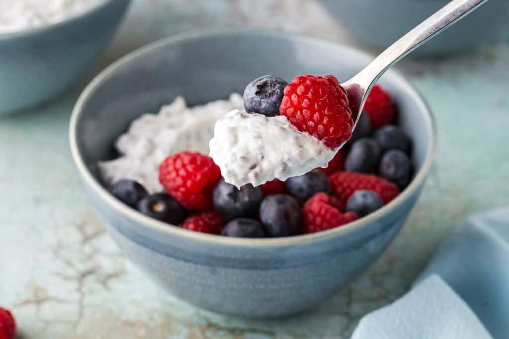 A low angle side shot of a high protein Greek yogurt bowl filled with fresh berries. A spoon has taken a scoop out. It is filled with creamy yogurt and chia seeds, and has a raspberry and blueberry too.
