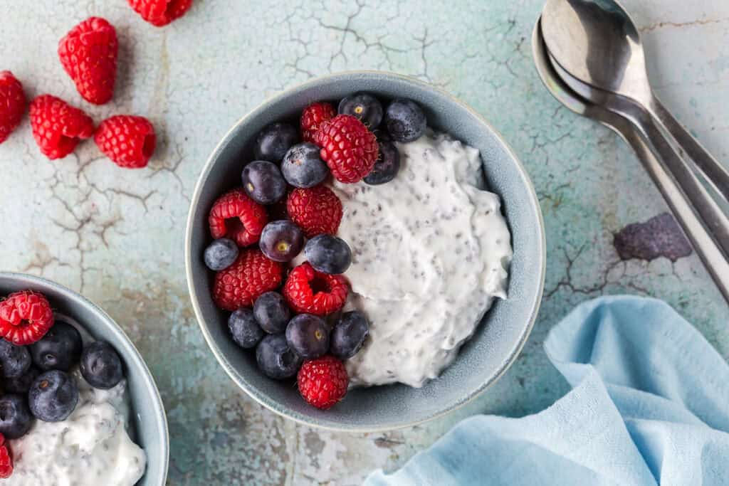 Overhead shot of a high protein Greek Yogurt bowl. Creamy Greek yogurt mixed with chia seeds and topped with fresh blueberries and raspberries. Fresh raspberries are scattered around the scene.