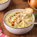 low angle side shot looking into a bowl of creamy clam chowder. A spoon is resting in the bowl and a chunk of rustic bread is dunked in the back of the bowl. Chunks of celery, carrots, clams, and bacon are present throughout.