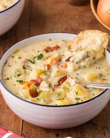 low angle side shot looking into a bowl of creamy clam chowder. A spoon is resting in the bowl and a chunk of rustic bread is dunked in the back of the bowl. Chunks of celery, carrots, clams, and bacon are present throughout.
