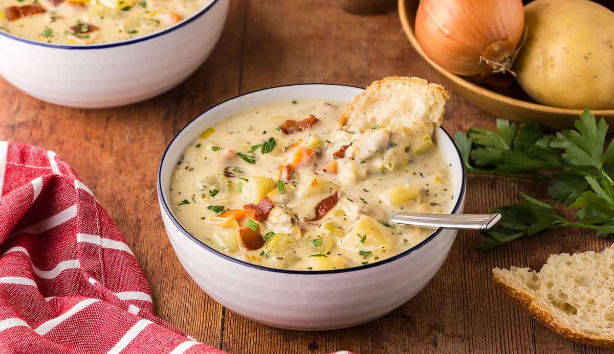 low angle side shot looking into a bowl of creamy clam chowder. A spoon is resting in the bowl and a chunk of rustic bread is dunked in the back of the bowl. Chunks of celery, carrots, clams, and bacon are present throughout.