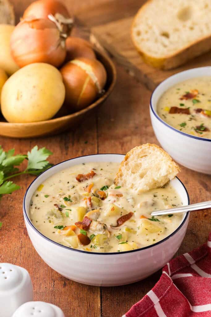 A vertical low angle shot peering into a white bowl of creamy clam chowder. A spoon is resting in the bowl and a chunk of rustic bread is dunked in the back of the bowl. Hearty chunks of celery, carrots, clams, and bacon are present throughout.  Another bowl of soup and fresh potatoes and onions are in the background.