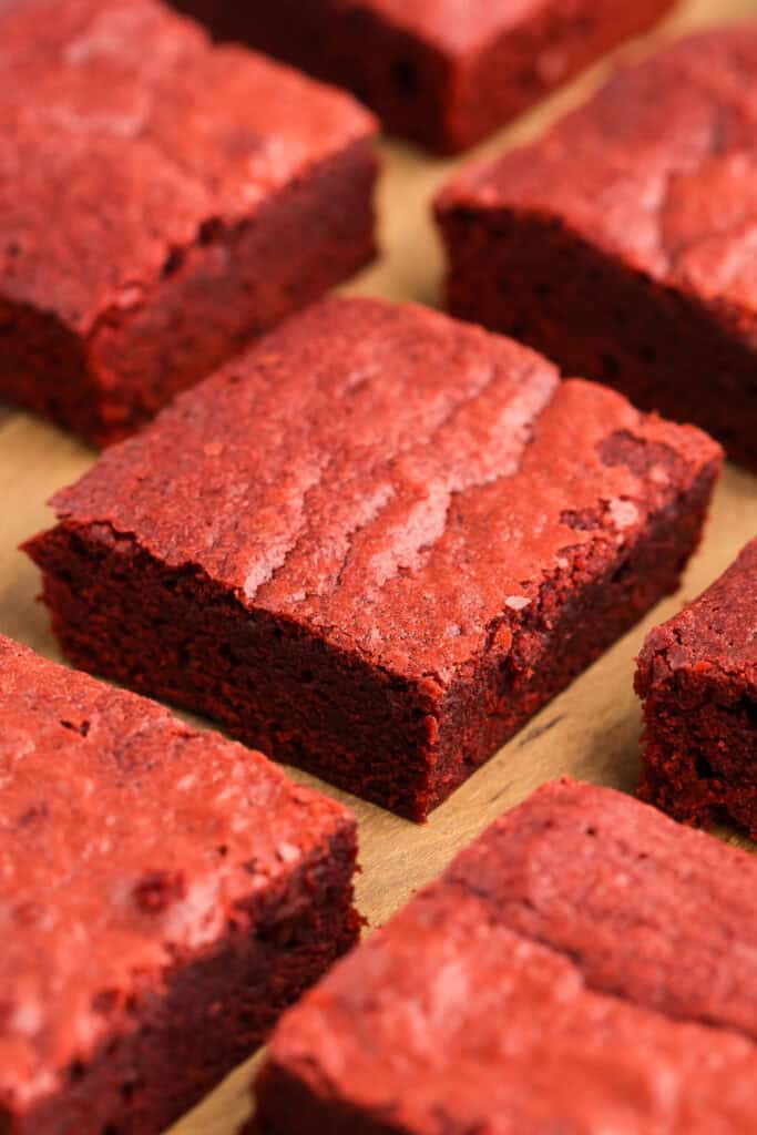 very close up low angle shot of a red velvet brownie in a sea of other red velvet brownies.  The top is shiny and crackly and the edges show a fudgy center.  All the brownies are a dark red hue and none have any frosting on top.