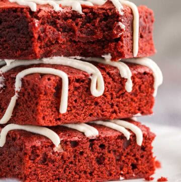 close up side shot of three red velvet brownies stacked on a small white plate. The top brownie has a bite taken out of it and is facing the camera showing off its fudgy center. All three brownies have been drizzled with a cream cheese frosting on top.