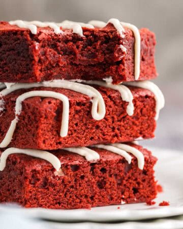 close up side shot of three red velvet brownies stacked on a small white plate. The top brownie has a bite taken out of it and is facing the camera showing off its fudgy center. All three brownies have been drizzled with a cream cheese frosting on top.
