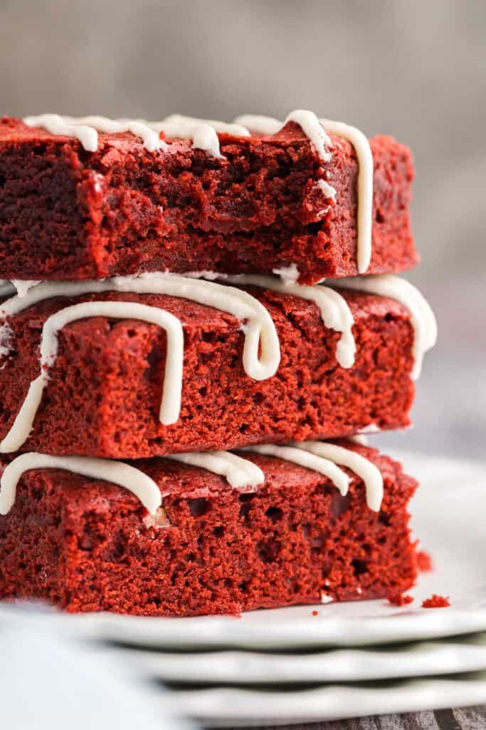close up side shot of three red velvet brownies stacked on a small white plate. The top brownie has a bite taken out of it and is facing the camera showing off its fudgy center. All three brownies have been drizzled with a cream cheese frosting on top.