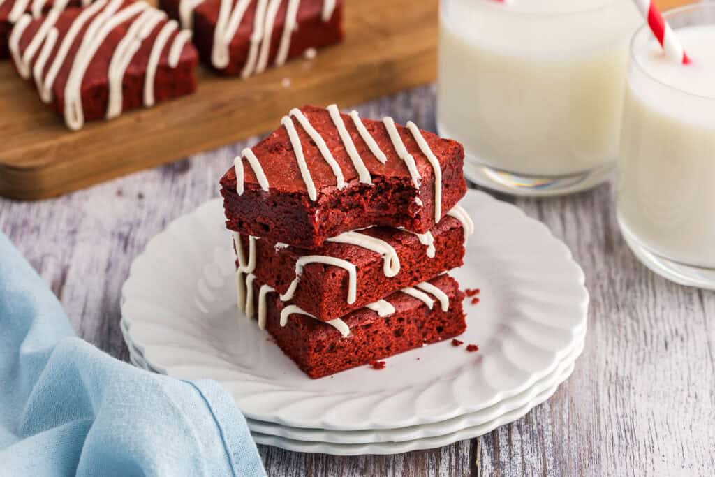 low angle shot of a stack of three red velvet brownies on a small white plate.  All brownies have a drizzle of cream cheese frosting and the top red velvet brownie has a bite taken out of it.  More brownies are in the background as well as two glasses of milk