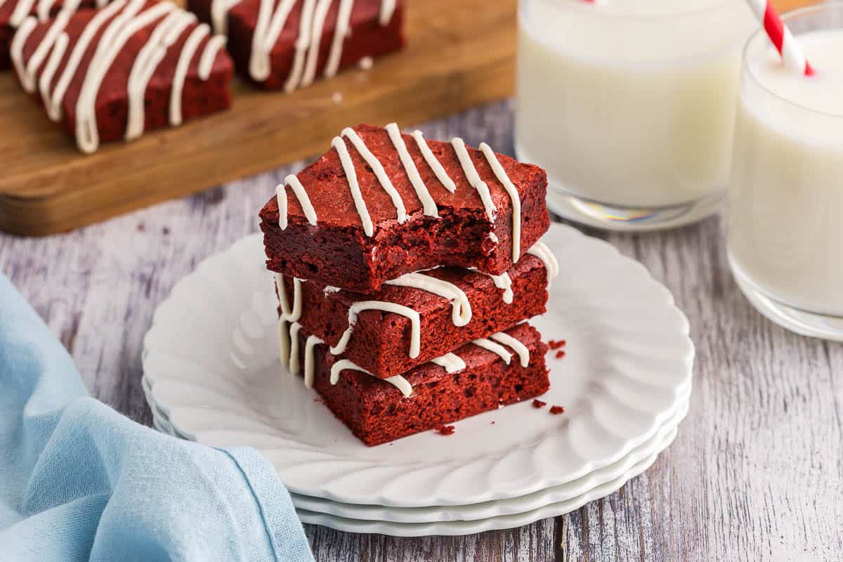 low angle shot of a stack of three red velvet brownies on a small white plate. All brownies have a drizzle of cream cheese frosting and the top red velvet brownie has a bite taken out of it. More brownies are in the background as well as two glasses of milk