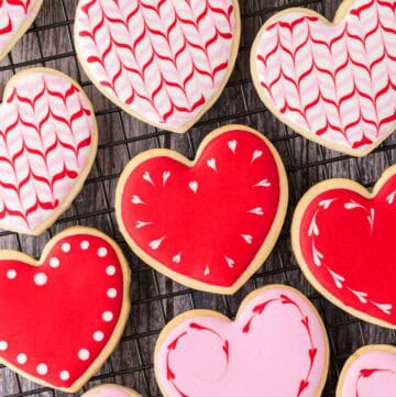 Overhead shot of heart-shaped sugar cookies on a cooling rack. Some are pink with a heart wreath in red around the edge. Some are red with white hearts around the edge, and some have a feathered white, red, and pink pattern.