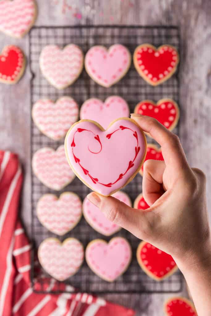 A hand is holding up a heart-shaped pink cookie close to the camera. Around the edge of the cookie are tiny red hearts. In the background is a cooling rack filled with more cookies.