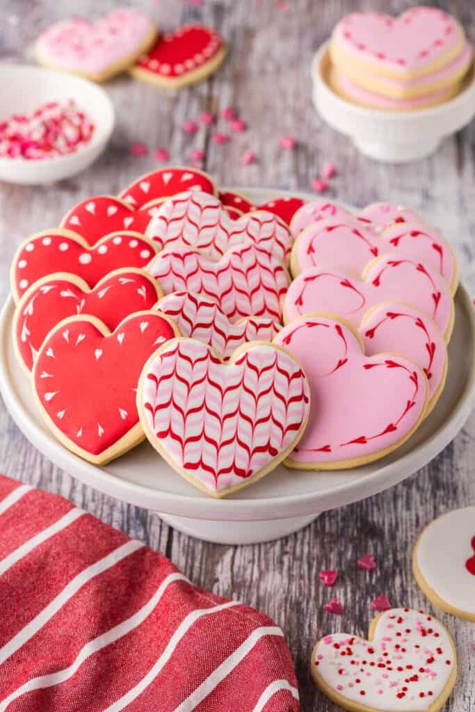 low angle shot of a batch of sugar cookies served on a white cake stand.  The sugar cookies have been cut into heart shapes and decorated with red, white, and pink icing for Valentine's Day.