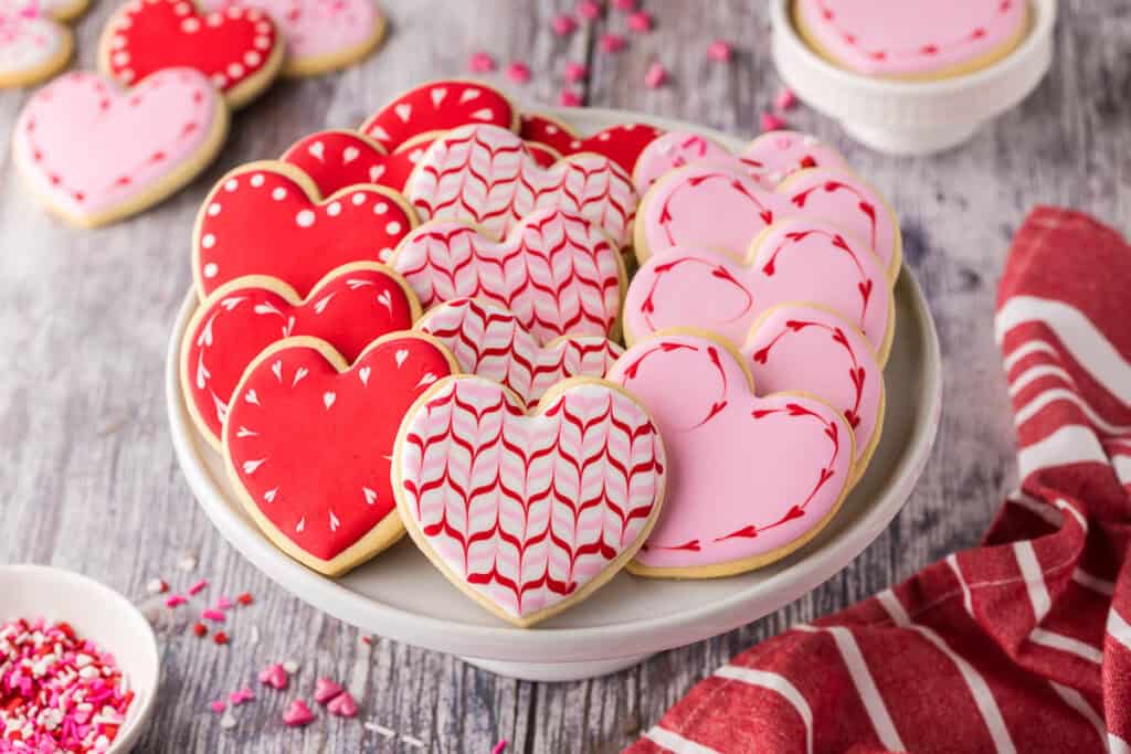horizontal orientation, low angle shot of a batch of sugar cookies served on a white cake stand.  The sugar cookies have been cut into heart shapes and decorated with red, white, and pink icing for Valentine's Day.