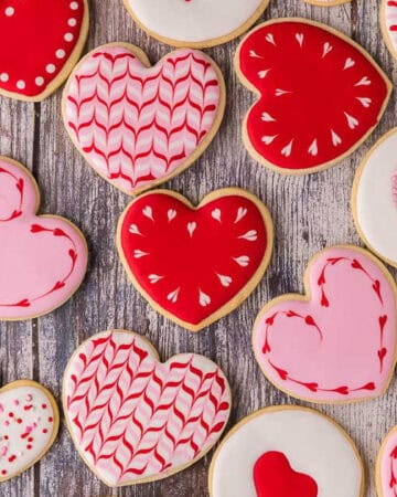 Overhead shot of various heart shaped sugar cookies. All cookies are piped with white, pink, or red royal icing. Some have sprinkles, some have heart-wreaths which have been scribed around the edges.