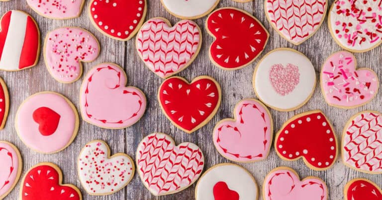 Overhead shot of various heart shaped sugar cookies. All cookies are piped with white, pink, or red royal icing. Some have sprinkles, some have heart-wreaths which have been scribed around the edges.
