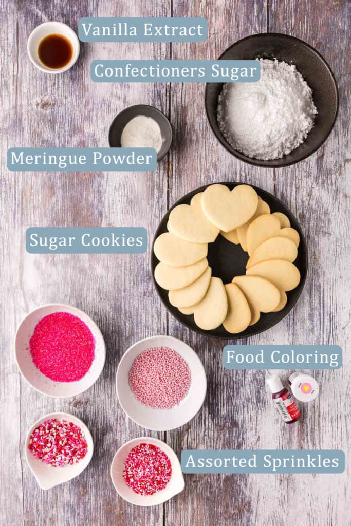 Overhead shot of the ingredients needed to decorate sugar cookies in a Valentine's theme. Ingredients include, sugar cookies, sprinkles, icing sugar, vanilla extract, and food coloring.