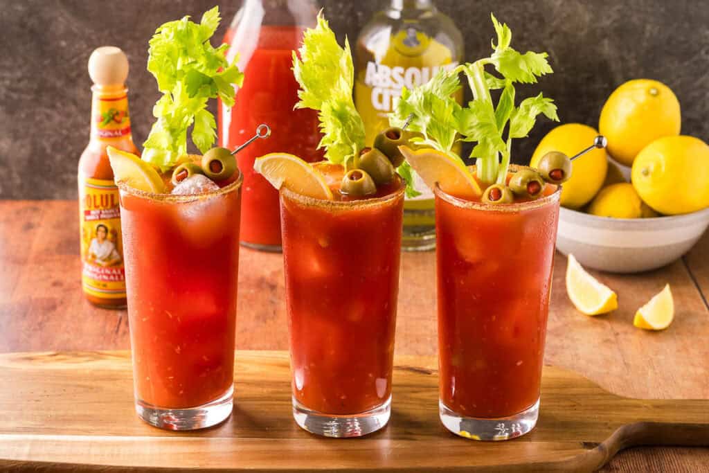 Three Bloody Marys on a wooden cutting board.  Each is garnished with a leafy celery stick, olives on a skewer, and a lemon wedge.  In the background is a bowl of lemons, as well as a vodka bottle and carafe of tomato juice.