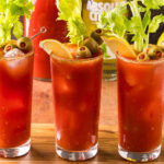 Three Bloody Marys on a wooden cutting board. Each is garnished with a leafy celery stick, olives on a skewer, and a lemon wedge. In the background is a bowl of lemons, as well as a vodka bottle and carafe of tomato juice.