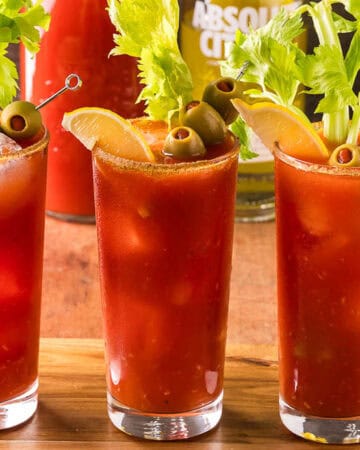 Three Bloody Marys on a wooden cutting board. Each is garnished with a leafy celery stick, olives on a skewer, and a lemon wedge. In the background is a bowl of lemons, as well as a vodka bottle and carafe of tomato juice.