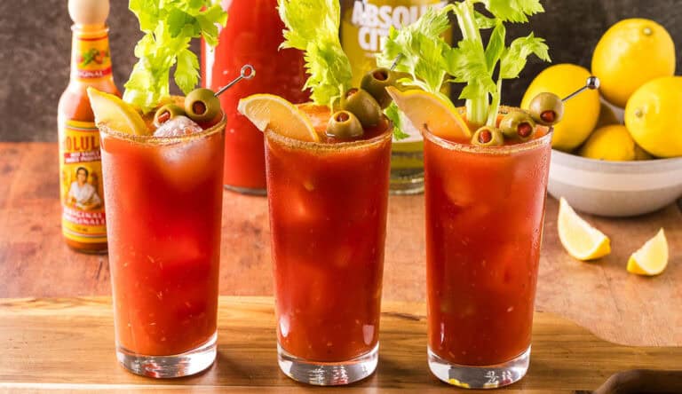 Three Bloody Marys on a wooden cutting board. Each is garnished with a leafy celery stick, olives on a skewer, and a lemon wedge. In the background is a bowl of lemons, as well as a vodka bottle and carafe of tomato juice.