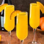 Three orange mimosas in champagne flutes garnished with an orange wedge on their rim. In the background is a bowl of oranges and a carafe or orange juice