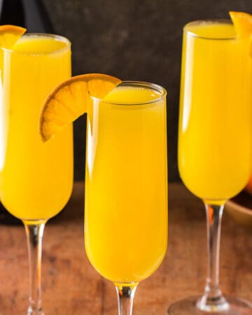Three orange mimosas in champagne flutes garnished with an orange wedge on their rim. In the background is a bowl of oranges and a carafe or orange juice