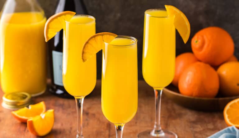Three orange mimosas in champagne flutes garnished with an orange wedge on their rim. In the background is a bowl of oranges and a carafe or orange juice