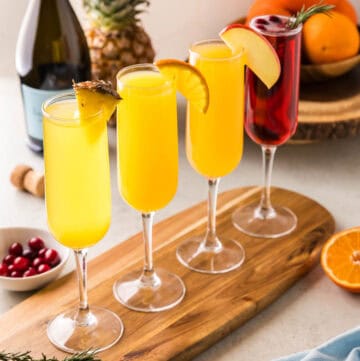 Side shot of 4 different flavors of mimosas in champagne flutes. The first is a pineapple mimosa garnished with a pineapple wedge. The next is an orange mimosa, garnished with an orange wedge. The third is an apple cider mimosa with an apple wedge, and last is a cranberry mimosa with cranberries and a sprig of rosemary.