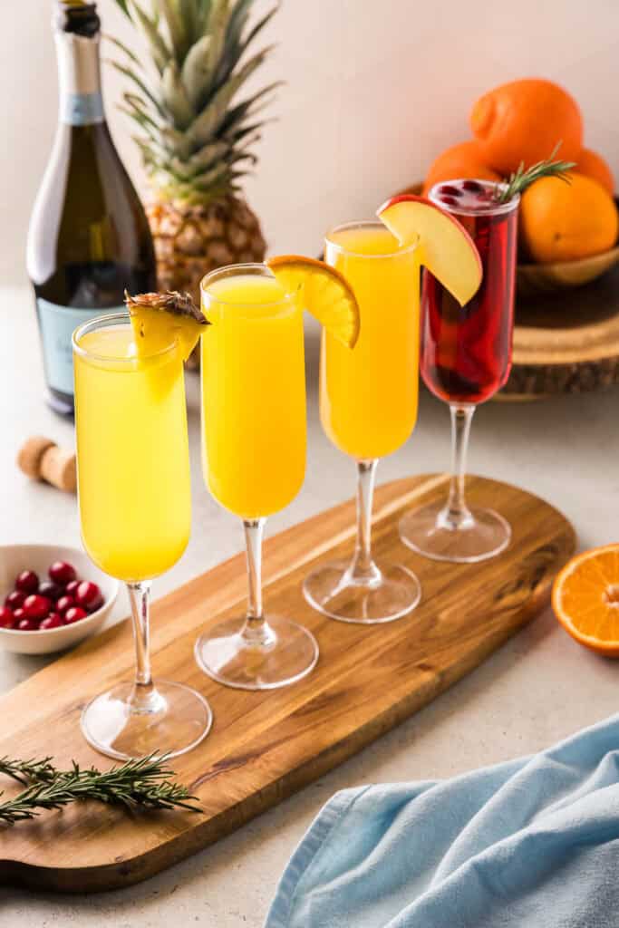 Side shot of 4 different flavors of mimosas in champagne flutes. The first is a pineapple mimosa garnished with a pineapple wedge. The next is an orange mimosa, garnished with an orange wedge. The third is an apple cider mimosa with an apple wedge, and last is a cranberry mimosa with cranberries and a sprig of rosemary.