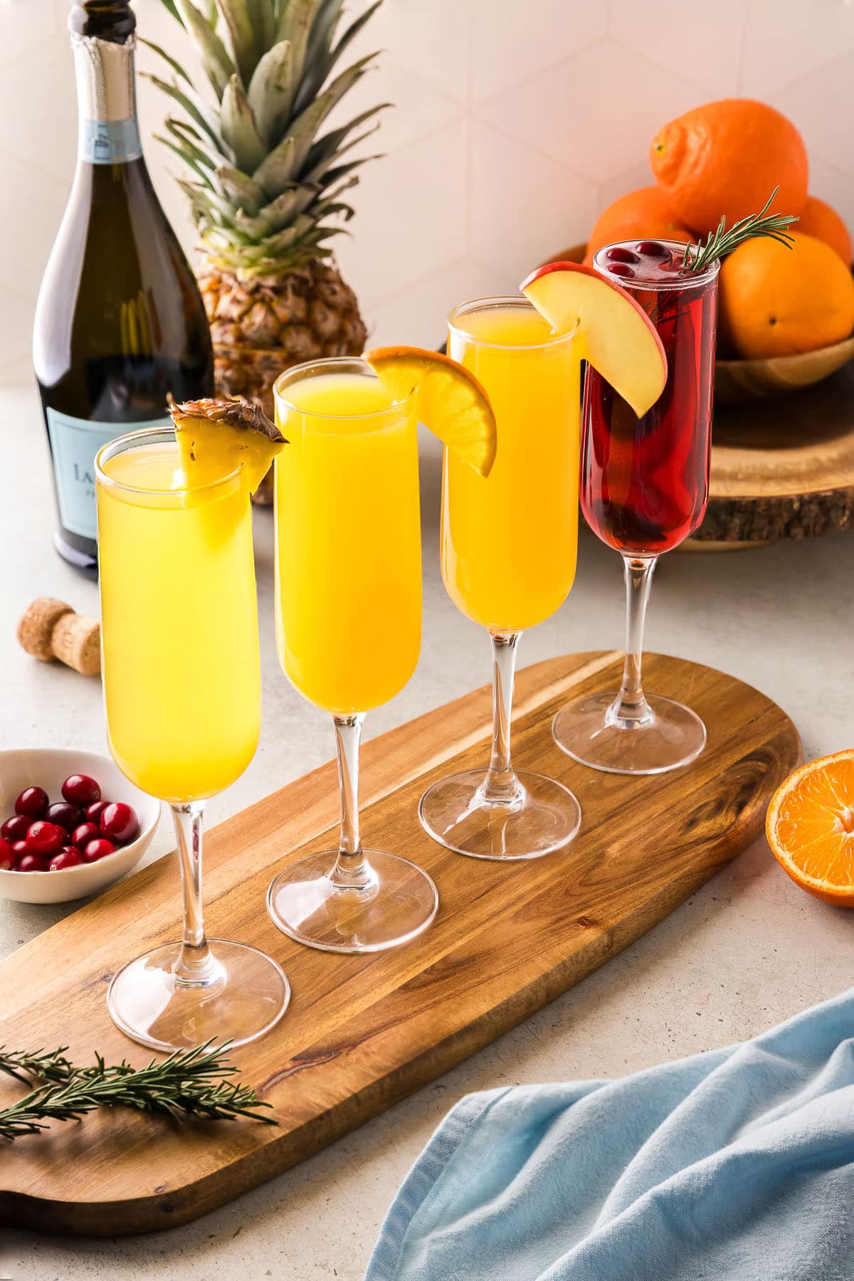 Side shot of 4 different flavors of mimosas in champagne flutes. The first is a pineapple mimosa garnished with a pineapple wedge. The next is an orange mimosa, garnished with an orange wedge. The third is an apple cider mimosa with an apple wedge, and last is a cranberry mimosa with cranberries and a sprig of rosemary.