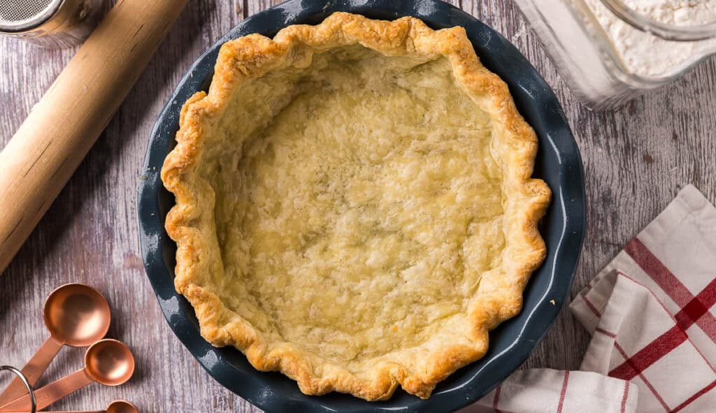 overhead shot of a par-baked golden brown pie crust in a blue ceramic pie plate. In the background there is a rolling pin, and measuring spoons.