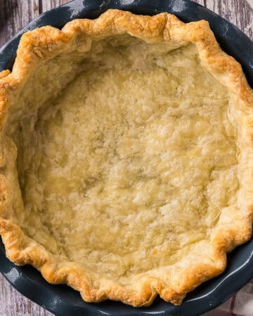 overhead shot of a par-baked golden brown pie crust in a blue ceramic pie plate. In the background there is a rolling pin, and measuring spoons.