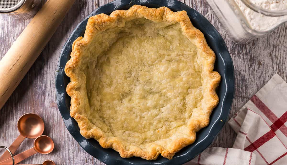 overhead shot of a par-baked golden brown pie crust in a blue ceramic pie plate. In the background there is a rolling pin, and measuring spoons.