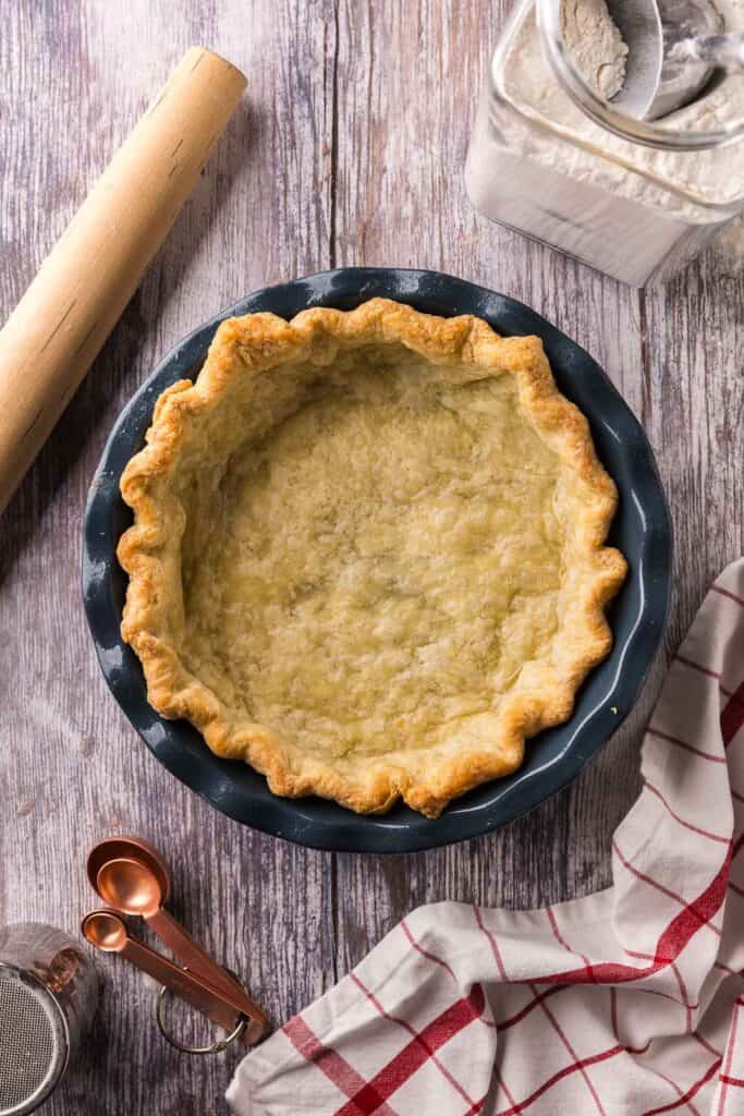 Overhead shot of a par-baked homemade pie crust. It is golden colored and sitting in a blue ceramic pie plate. There is a rolling pin, copper measuring spoons, and a canister of flour in the scene.