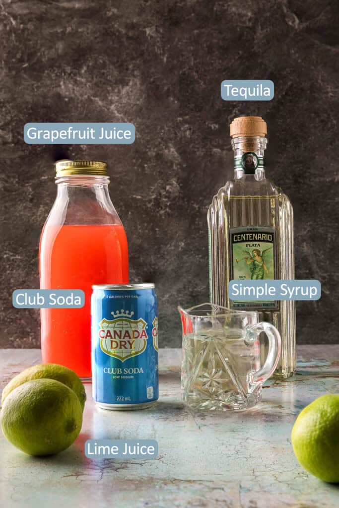 side shot of the ingredients used to make this paloma cocktail recipe.  Tequila, grapefruit juice, limes, club soda, and simple syrup.