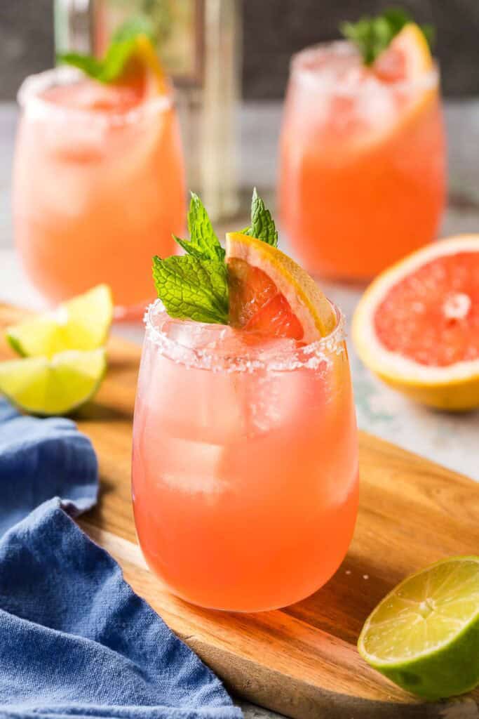 A low angle 45 shot of a non-stemmed wine glass filled with a pink paloma cocktail.  In the background are two more palomas.  All are garnished with a grapefruit wedge and a sprig of mint in the glass.