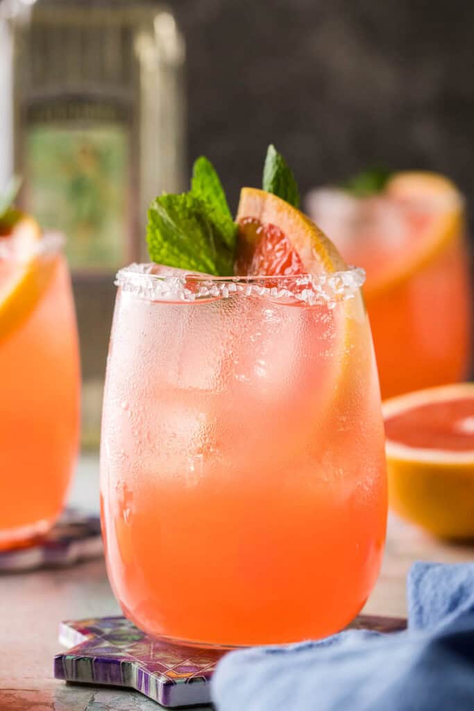 Close up side shot of a paloma drink.  This pink cocktail is garnished with a grapefruit wedge and a sprig of mint.