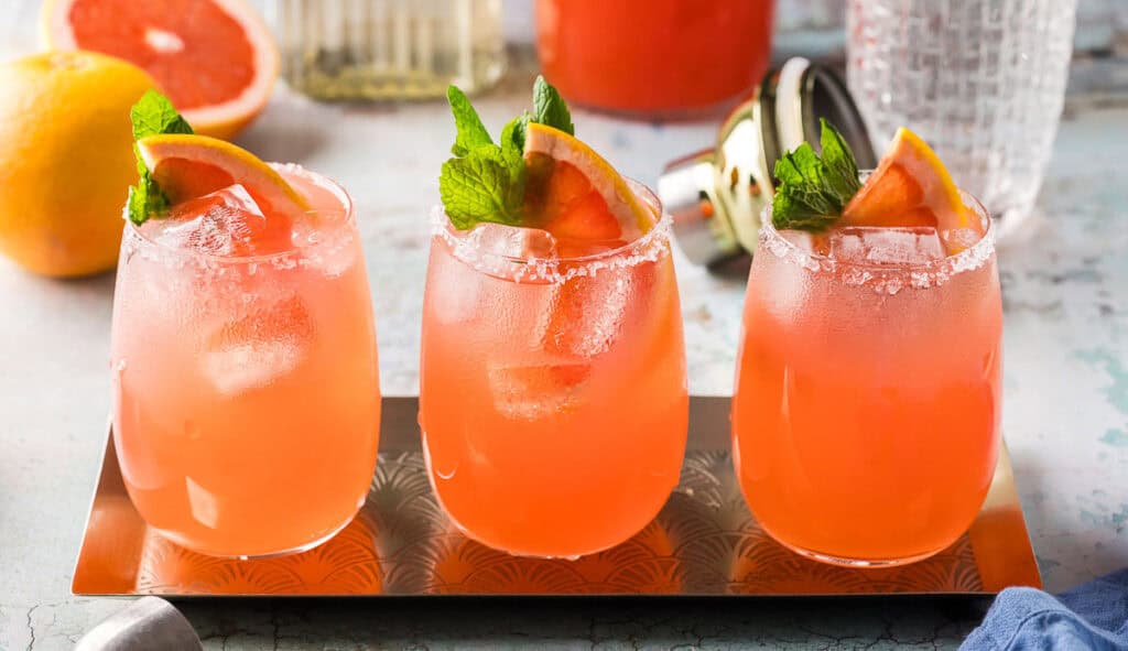 Three light pink paloma cocktails side by side on a stainless steel rectangular serving tray. Each cocktail is garnished with a grapefruit wedge and sprig of mint.
