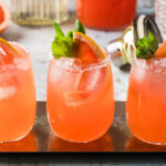 Three light pink paloma cocktails side by side on a stainless steel rectangular serving tray. Each cocktail is garnished with a grapefruit wedge and sprig of mint.