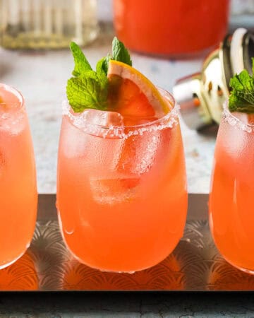 Three light pink paloma cocktails side by side on a stainless steel rectangular serving tray. Each cocktail is garnished with a grapefruit wedge and sprig of mint.