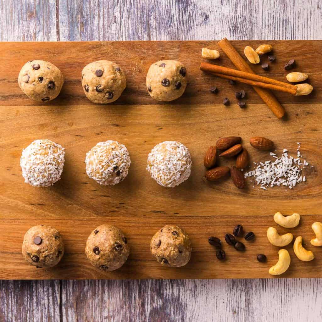 Overhead shot of a wooden cutting board with three lines of protein balls.  The top line of three balls are peanut butter protein balls, the second line of three balls is cashew coconut protein balls, and the final line is almond espresso protein balls.  