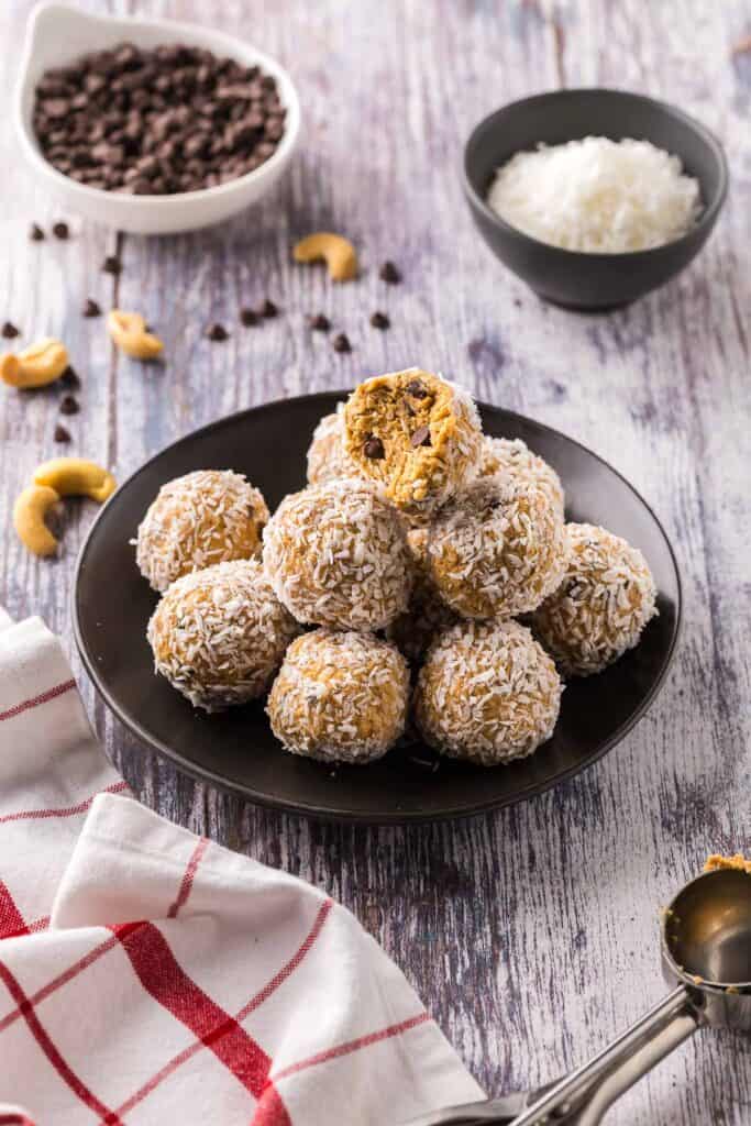 Side shot of a small black plate filled with coconut protein balls.  The top ball has a bite taken out of it.  All balls contain chocolate chips and have been rolled in shredded coconut.