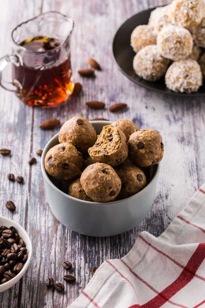 A small baby blue bowl filled with almond espresso protein balls.  The top one has a bite out of it.  All balls contain chocolate chips.  In the background is a small jar of maple syrup and plate filled with coconut protein balls.