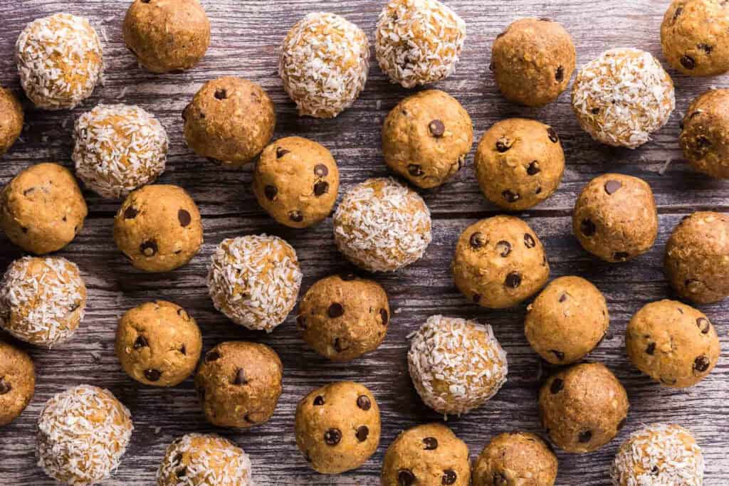 A horizontal close-up overhead shot of three flavors of protein balls all randomly filling up the space on a wooden table.  Some are coated in coconut, and most have chocolate chips.