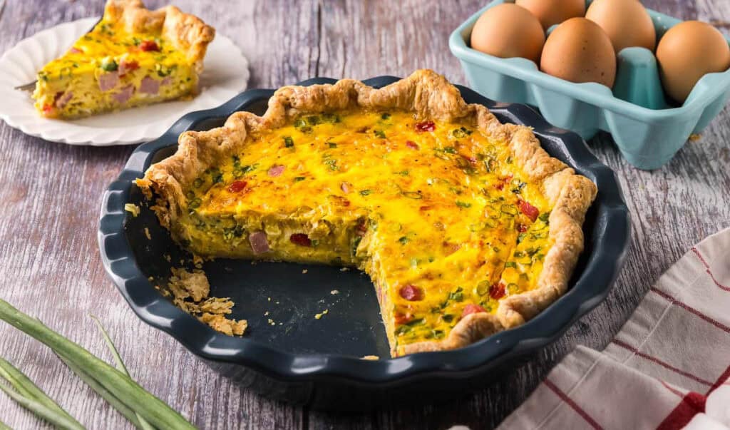 horizontal picture of a blue pie plate with a golden yellow quiche inside. Two slices have been removed revealing a creamy custard studded with ham, red peppers, and onion chunks. The crust is rustic and homemade.