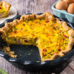 horizontal picture of a blue pie plate with a golden yellow quiche inside. Two slices have been removed revealing a creamy custard studded with ham, red peppers, and onion chunks. The crust is rustic and homemade.