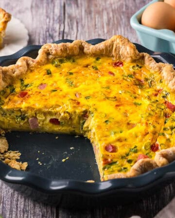 horizontal picture of a blue pie plate with a golden yellow quiche inside. Two slices have been removed revealing a creamy custard studded with ham, red peppers, and onion chunks. The crust is rustic and homemade.