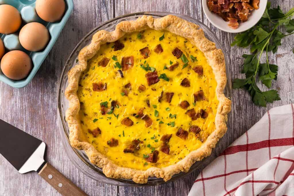 A quiche Lorraine in a glass pyrex pie plate made with a homemade rustic crust. Bacon, parsley, eggs, and a pie server are all in the background.