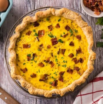 A quiche Lorraine in a glass pyrex pie plate made with a homemade rustic crust. Bacon, parsley, eggs, and a pie server are all in the background.